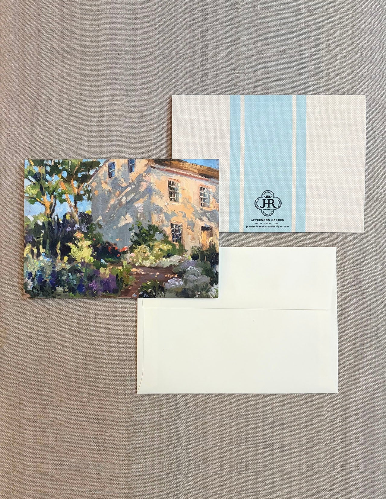 A stationery set laid flat on a textured beige fabric background, including a printed card with a painted house and garden scene, a white envelope, and a white card with pale blue vertical stripes and a small monogram logo.