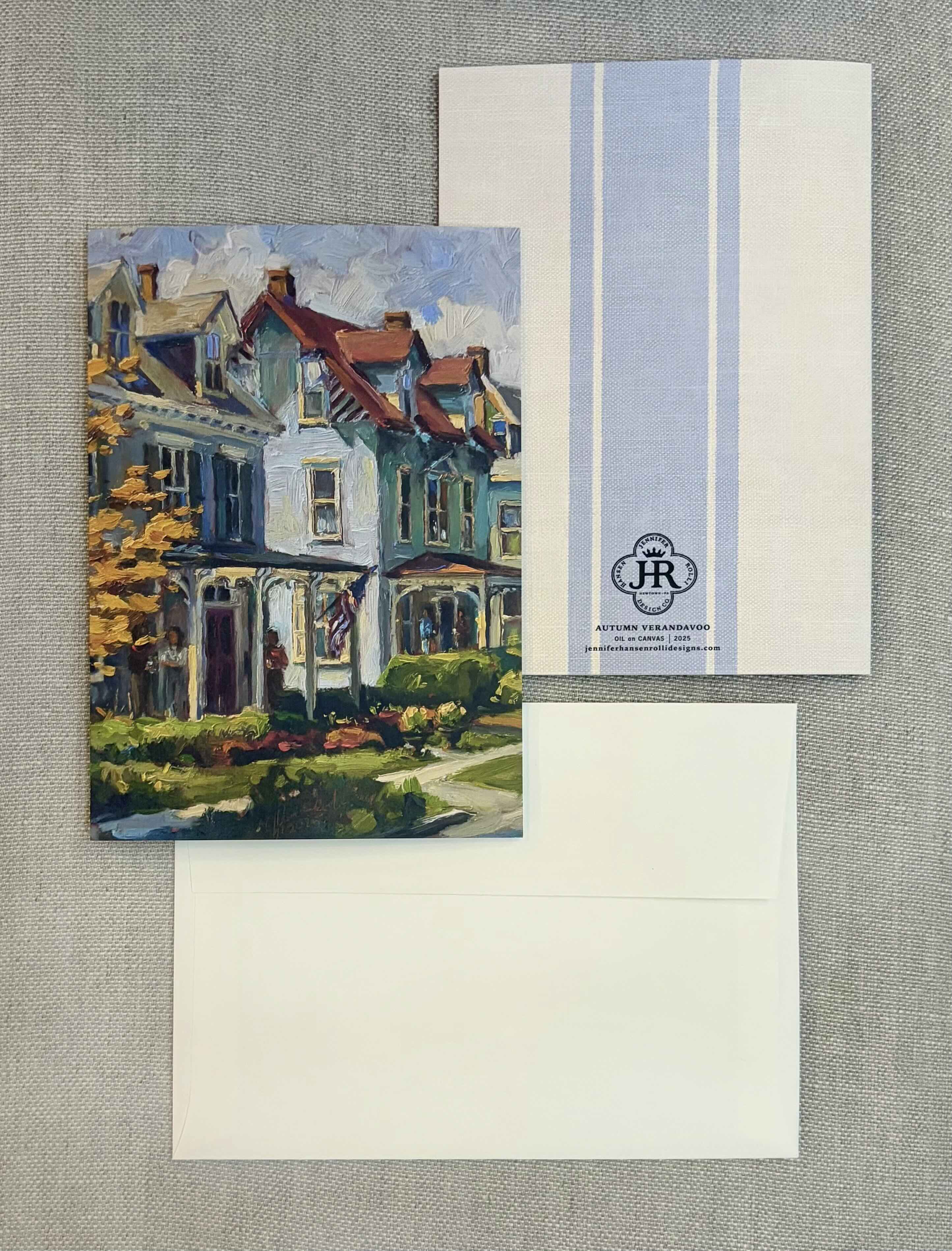 A stationery set laid flat on a textured gray fabric background, featuring a printed card with a painted house illustration, a white envelope, and a white card with pale blue vertical stripes and a small monogram logo.