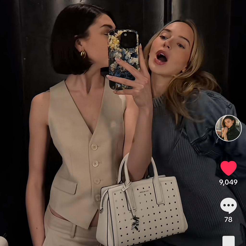 Two women taking a mirror selfie with a phone, one holding a handbag.