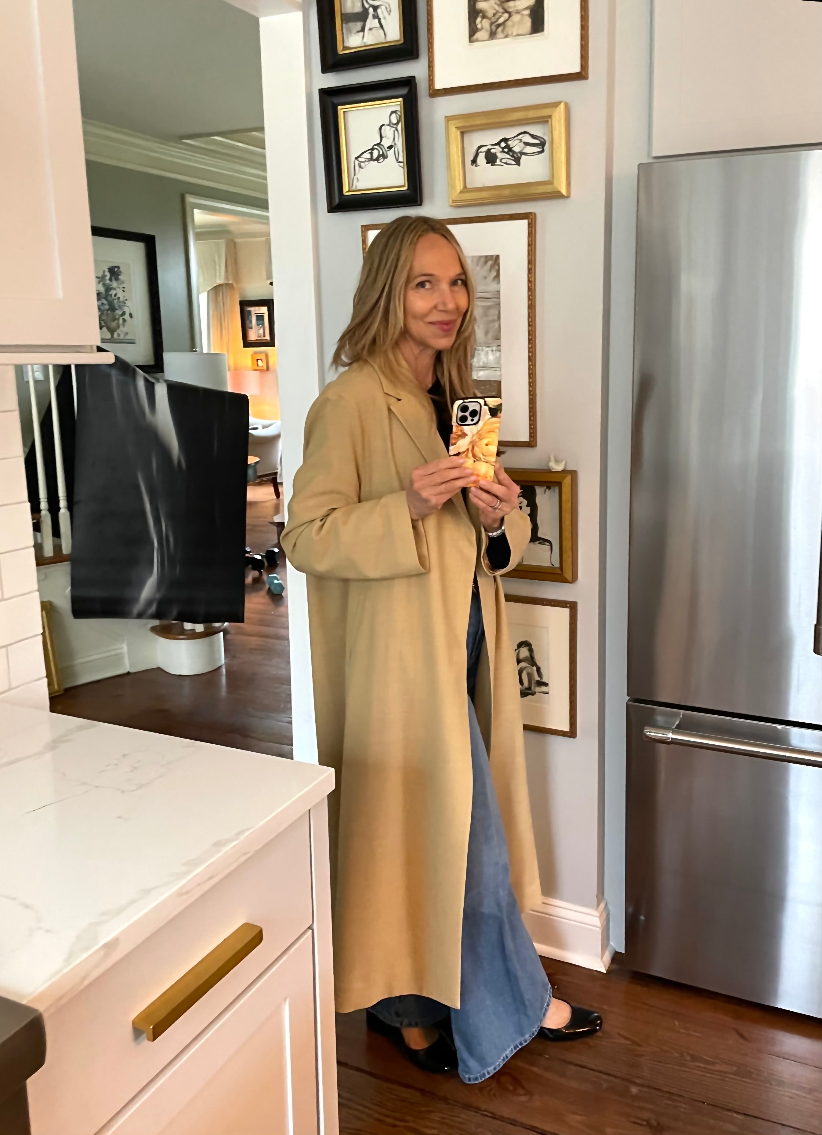 Woman in a long beige coat standing in a kitchen holding a phone.