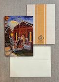 A stationery set laid flat on a textured gray fabric background, including a printed card with an impressionistic painting of a building exterior at dusk, a white envelope, and a white card with two vertical gold stripes and a small monogram logo.