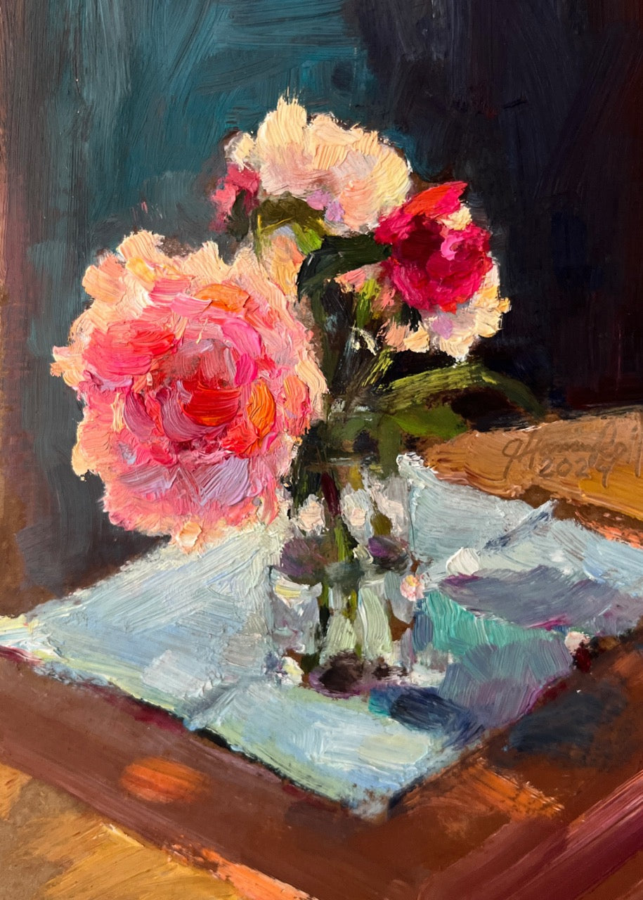 An oil painting of a bouquet of pink, peach, and red flowers in a clear glass vase resting on a light blue cloth over a brown wooden table, set against a dark teal and black background.