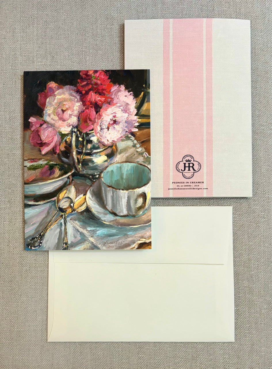 A stationery set featuring a printed card with an oil painting of pink flowers and a teacup, shown with its matching envelope and backing card on a textured gray fabric background.
