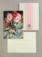 A stationery set featuring a printed card with an oil painting of pink flowers and a teacup, shown with its matching envelope and backing card on a textured gray fabric background.