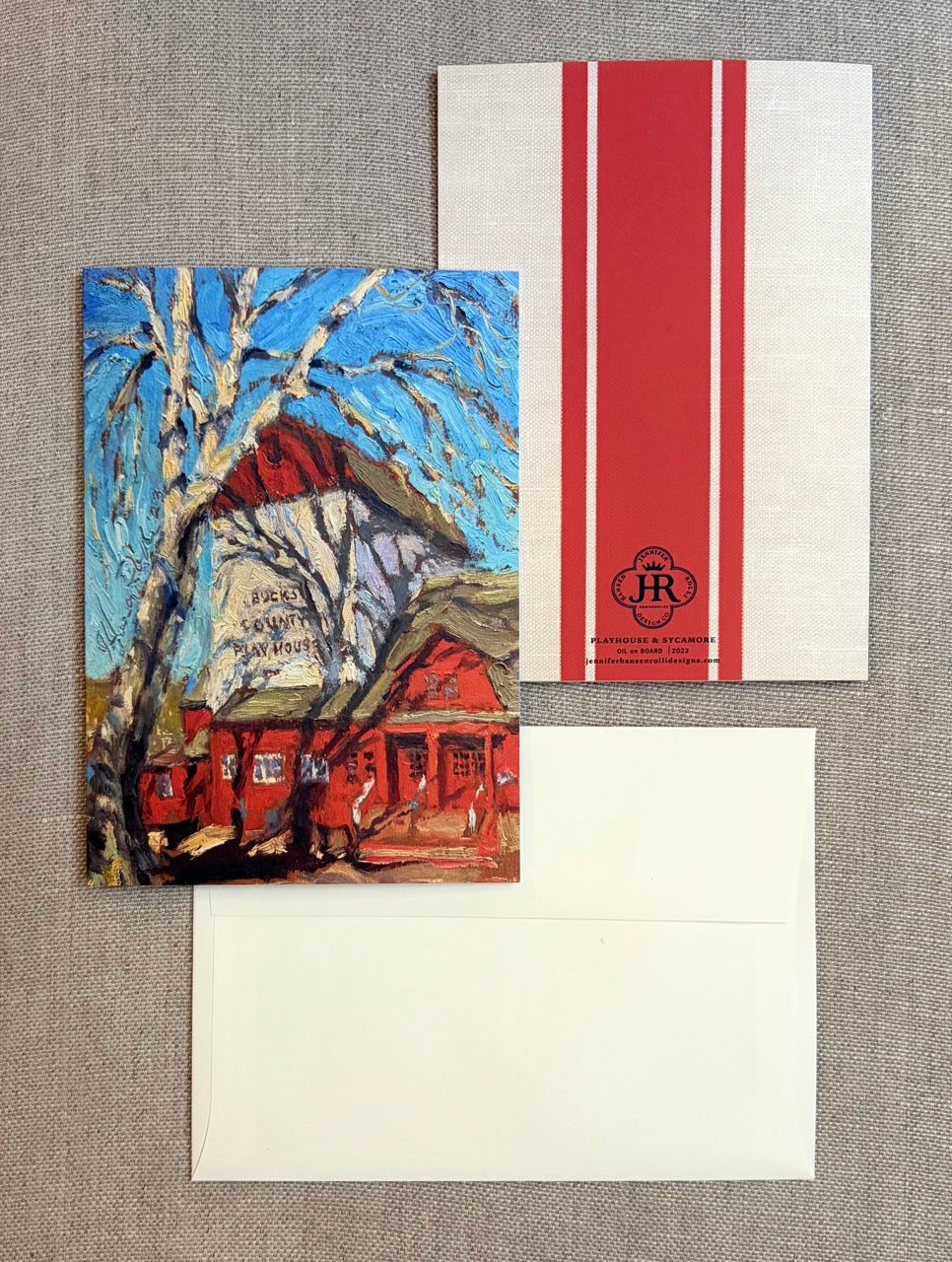 A stationery set laid flat on a textured beige fabric background, including a printed greeting card with an impressionistic red house and blue sky scene, a white envelope with a bold red vertical stripe and a small circular 'JR' logo, and another white envelope below.