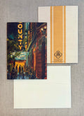 A stationery set laid flat on a textured gray fabric background, featuring a printed card with an impressionistic street scene and a vertical gold stripe, a matching cream envelope, and another cream envelope behind it. The printed card has a gold 'COUNTY' sign and a circular 'JR' monogram logo.