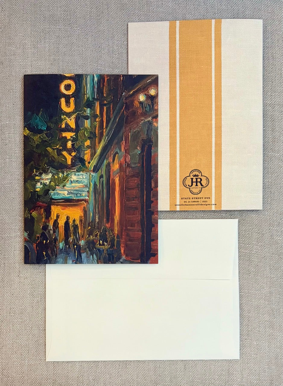 A stationery set laid flat on a textured gray fabric background, featuring a printed card with an impressionistic street scene and a vertical gold stripe, a matching cream envelope, and another cream envelope behind it. The printed card has a gold 'COUNTY' sign and a circular 'JR' monogram logo.